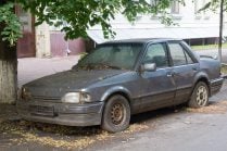 Home - Auto Wreckers A dirty, abandoned grey sedan parked on a street under a tree, covered in leaves and debris.