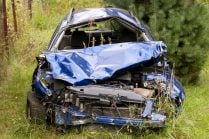 Home - Auto Wreckers A severely wrecked blue car with a crushed front end and hood, abandoned in a grassy, overgrown field near some trees.