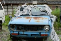 Home - Auto Wreckers Front view of an old, rusty light-blue car with a missing grille and headlight, partially covered by a shredded grey tarp in a grassy yard.